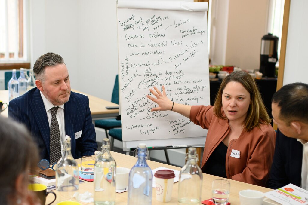 A group of smartly-dressed people sit around a table filled with notebooks, water bottles and coffee cups. One of the women is speaking and pointing towards a flipchart, which is covered in black writing.