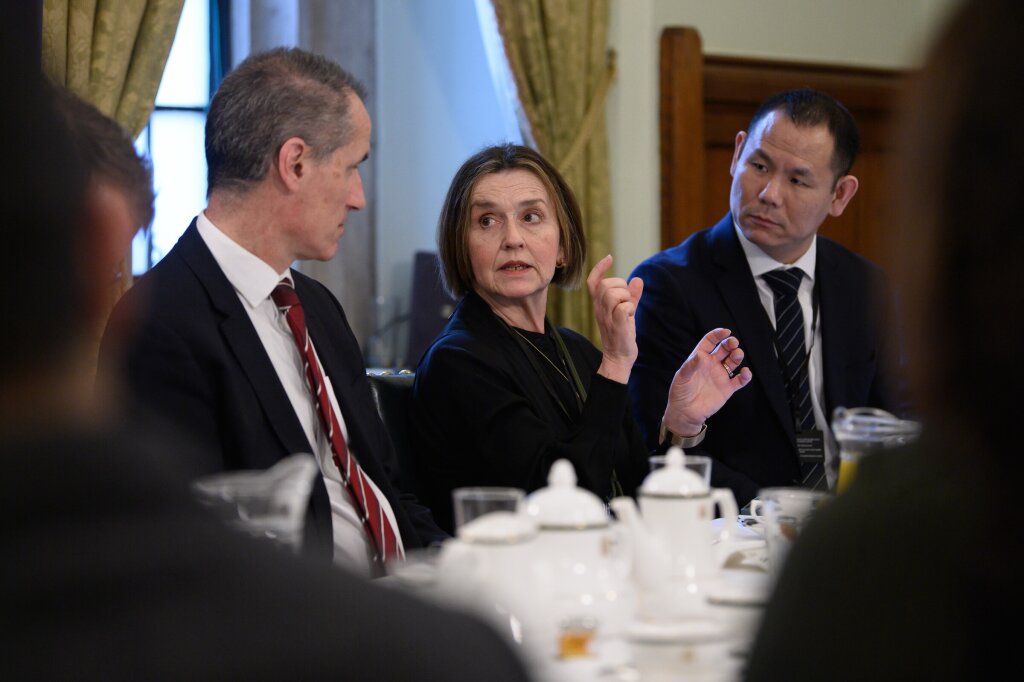 A woman sits in between two men dressed in suits at a table filled with white crockery. The woman is speaking using her hands, and turning towards the man on her left.