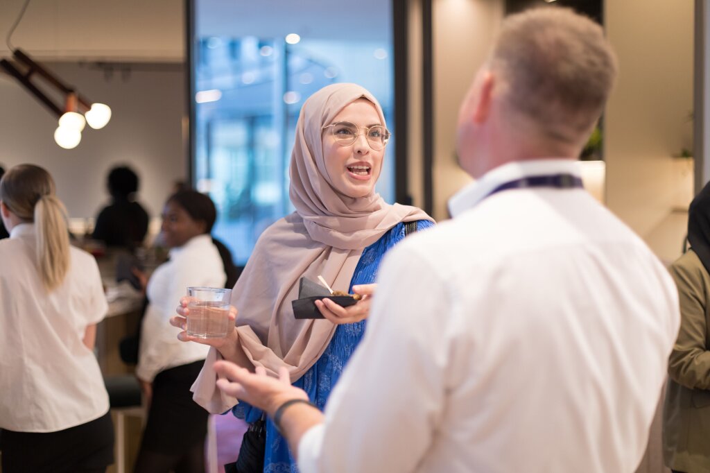 A woman in a blue dress and a beige hijab speaks to a man in a white shirt who's back is turned from the camera.