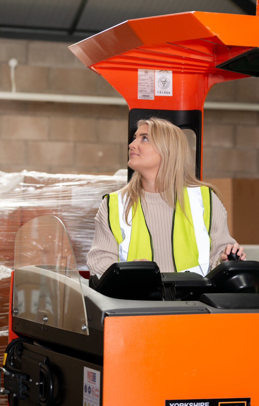 A woman with long blonde hair and hi viz vest driving a piece of machinery.