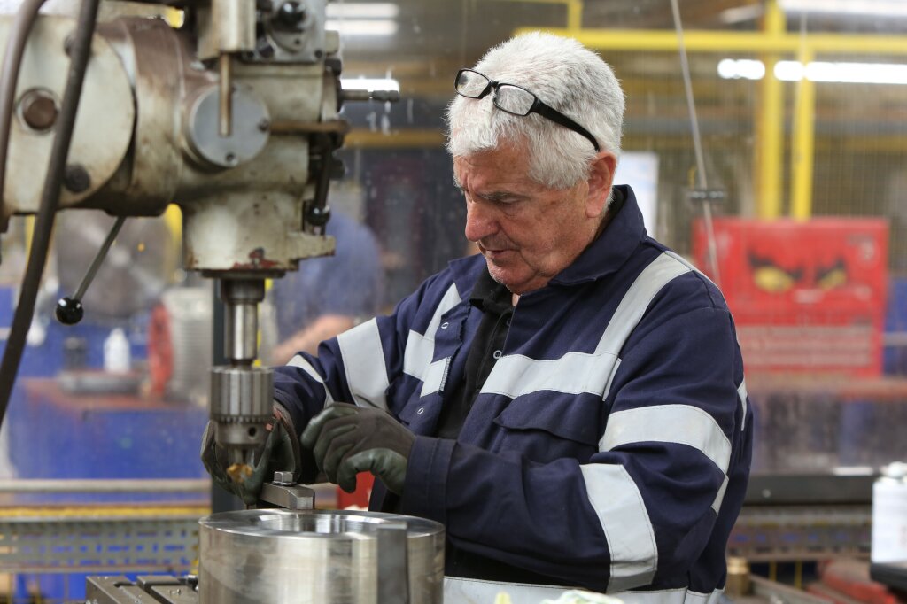 A man with short white hair works on a large piece of machinery in a factory. His black glasses are perched on the top of his head and he is wearing blue overalls with hi-viz strips and dark green gloves.