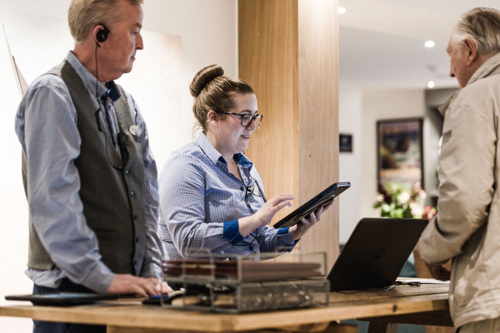 A man and a woman, both dressed in blue check shirts, work at the front desk of a hotel. The woman is checking a male customer into the hotel using a handheld tablet.
