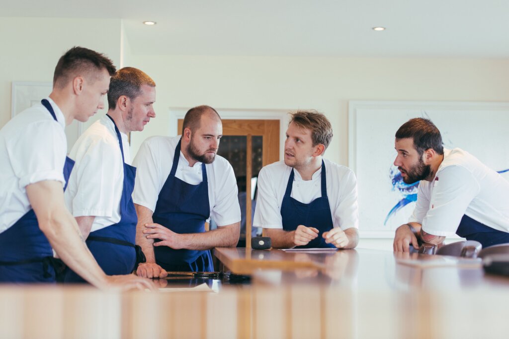 Five male chefs in chef's whites and blue aprons gather around a brown table for a meeting.