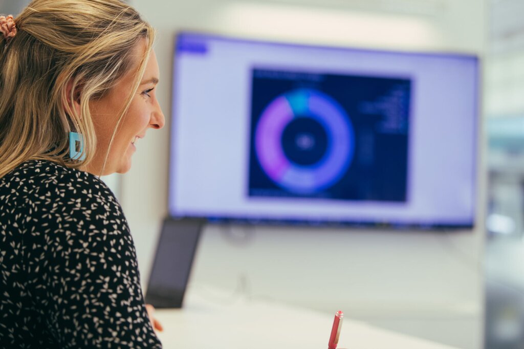 A woman with blonde hair, blue earrings and a patterned black shirts sits in front of a large TV screen showing a pie chart. The graph is out of focus.