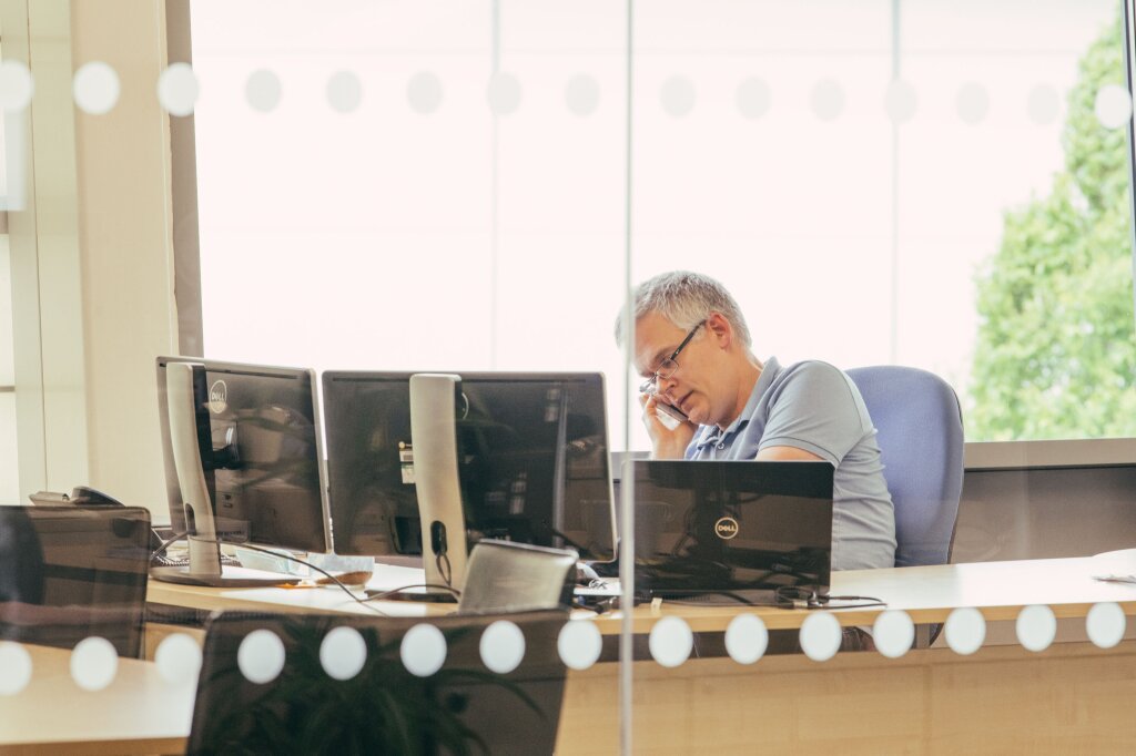 A man in a grey t-shirt works in an office with glass walls. He is facing two desktop displays and an open laptop and is speaking on a mobile phone.