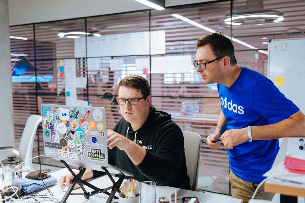 Two men, both wearing glasses, work together on a laptop, the back of which is covered in stickers.