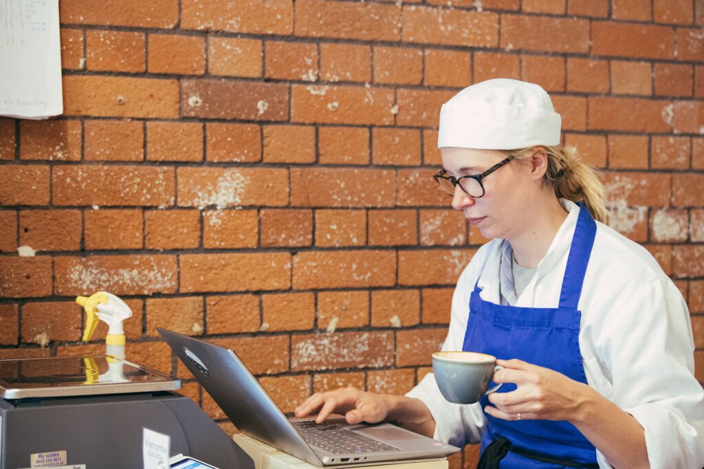 A female chef in chef's whites a blue apron sits at a laptop. She is typing with one hand and holding a cup of coffee with the other.